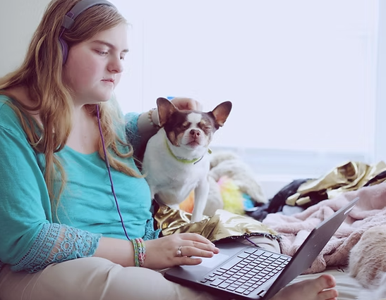 Young Woman Sitting In Her Bed, Holding Her Dog While On The Laptop